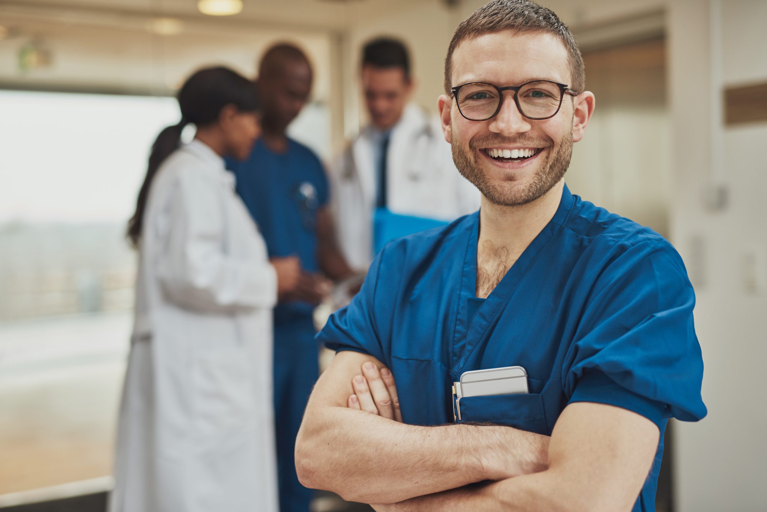 Smiling optimistic young male hospital surgeon standing in front of his colleagues with folded arms beaming at the camera